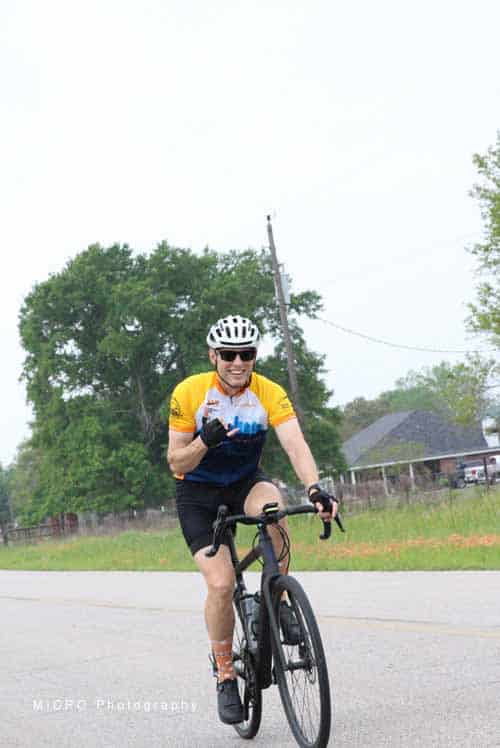 Bright cyclist riding Orange Bluebonnet Express bicycle in open countryside with greenery and rural houses.