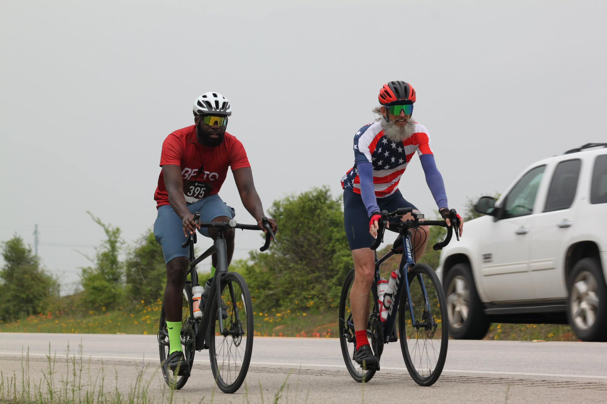 Vibrant cyclists riding on the road with greenery and a white vehicle in the background.