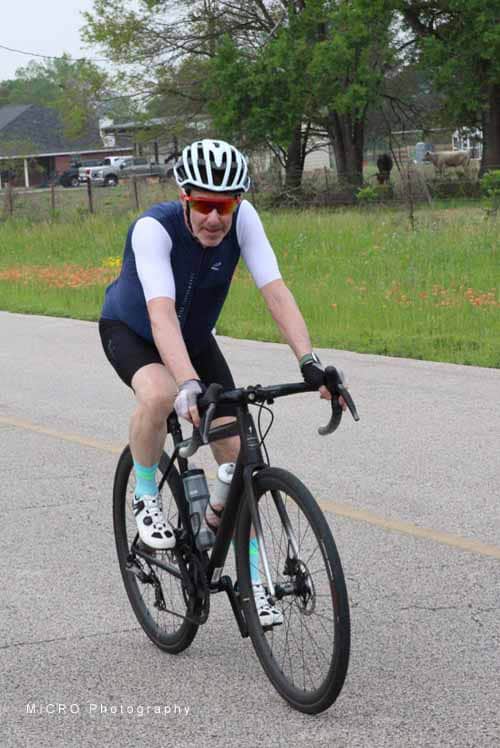 Bright male cyclist wearing helmet and sunglasses riding a black road bike on a paved road outdoors.
