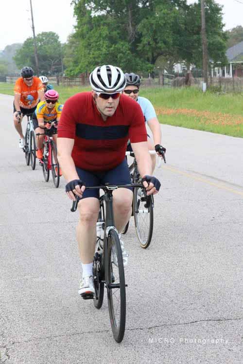 Brightly colored group cycling on suburban road during daytime, wearing helmets and athletic clothing.