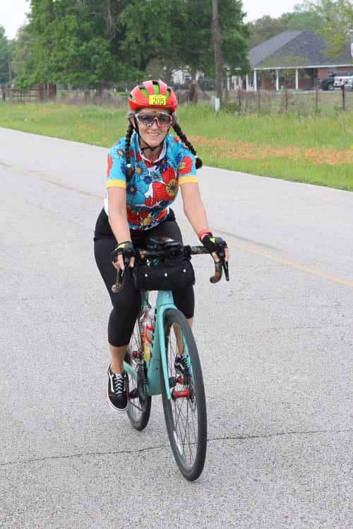 Colorful female cyclist wearing a vibrant floral jersey, helmet, and sunglasses riding a bike outdoors.