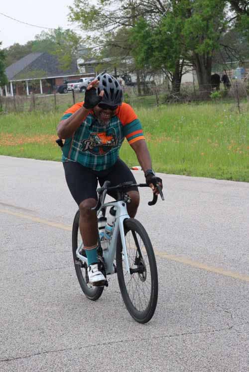 Man riding bicycle on rural road, smiling and waving, enjoying cycling event; Bluebonnet Express cycling race.
