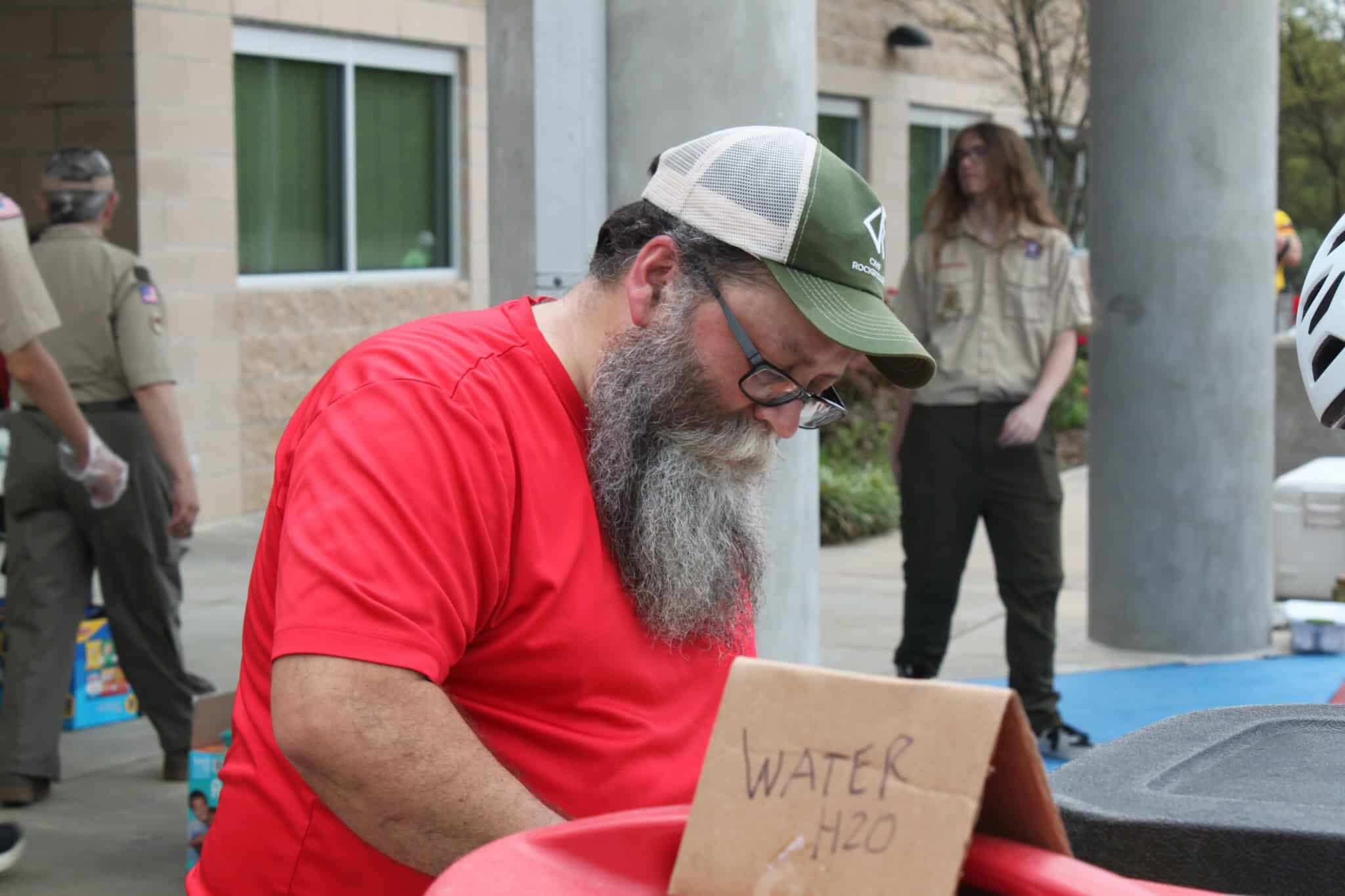 Man with a beard and glasses wearing a red shirt and green hat at outdoor event.
