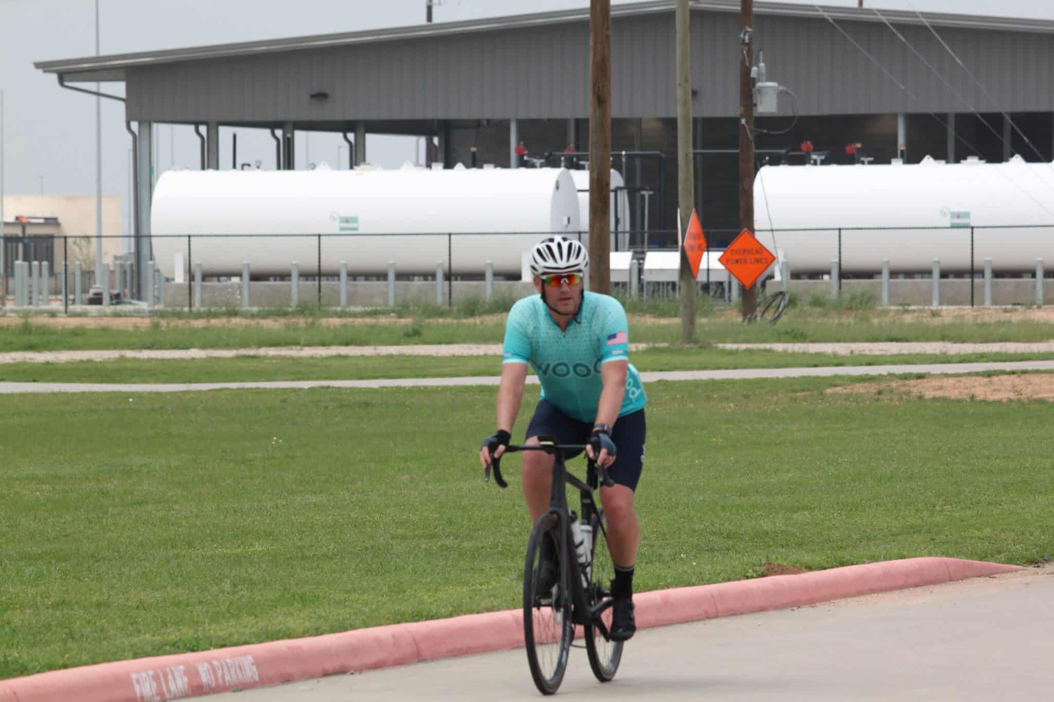 Bright blue cyclist jersey riding a bike on a paved path with fuel storage tanks in background.
