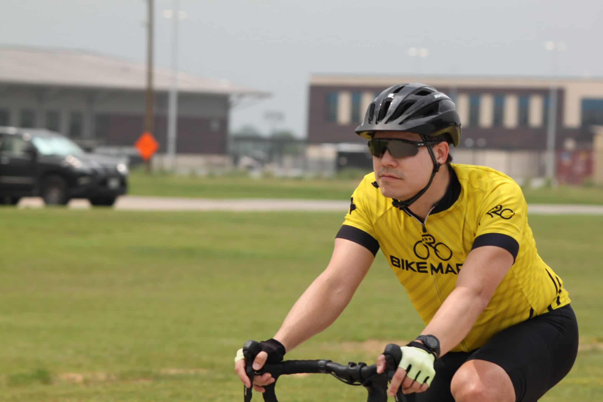 Bike courier riding bicyclist on city street with BikeMart logo, yellow cycling jersey, black helmet, and sunglasses.