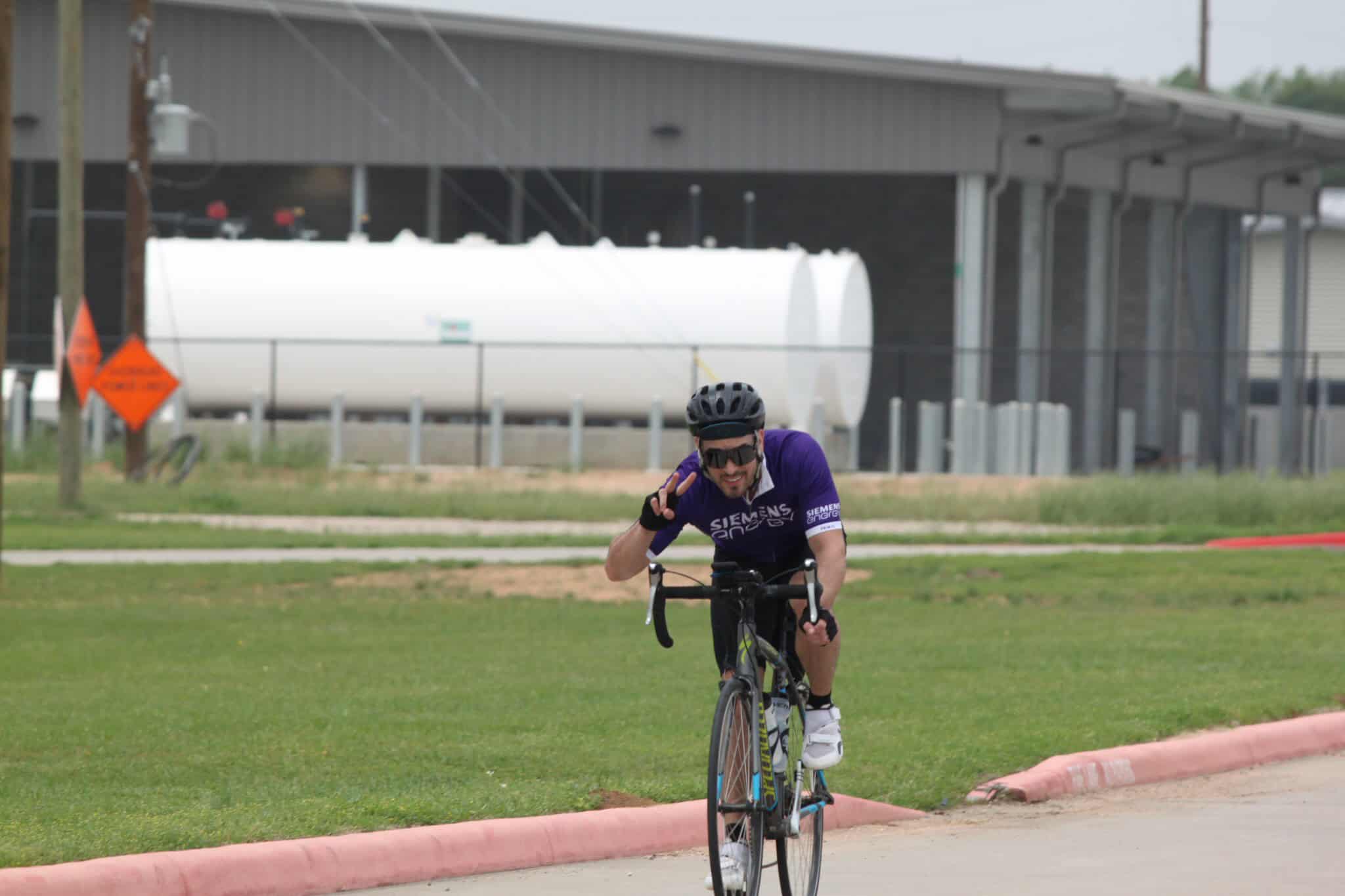 Rider wearing purple cycling jersey, helmet, and sunglasses, riding a bike during a cycling event.