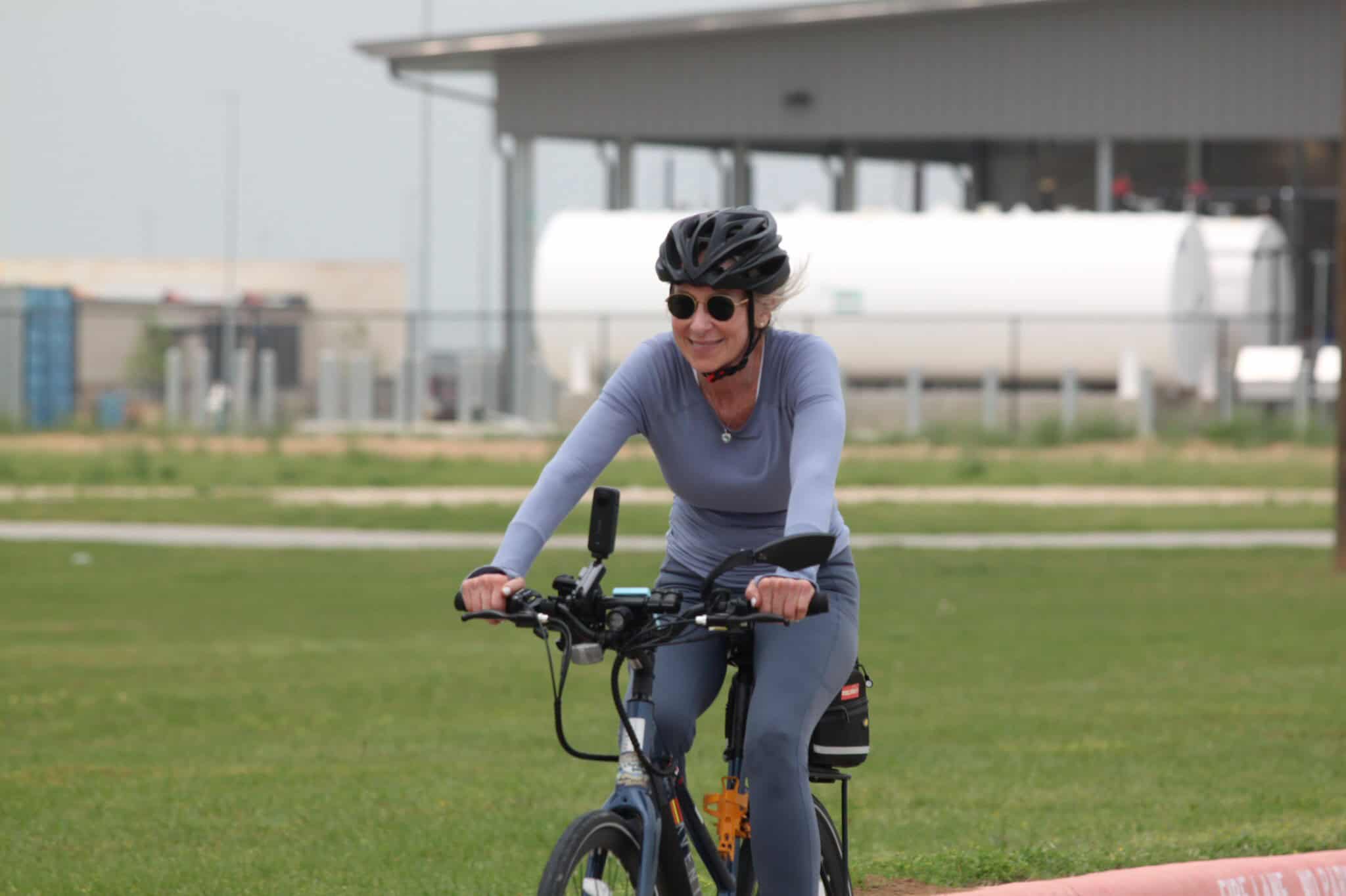Women riding electric bike on grassy trail, smiling, wearing helmet and sunglasses, eco-friendly transportation, Bluebonnet Express.