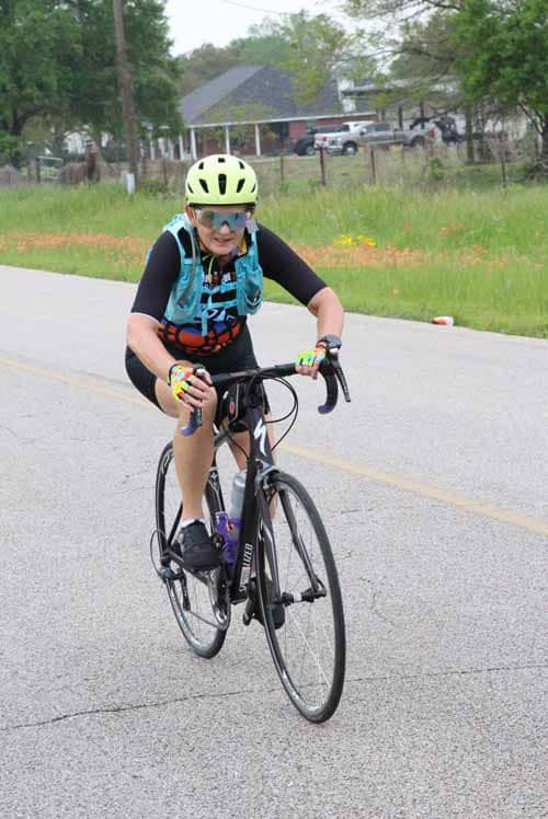 Bright female cyclist riding a black bike on a rural road during daytime, wearing safety gear and a blue jersey.