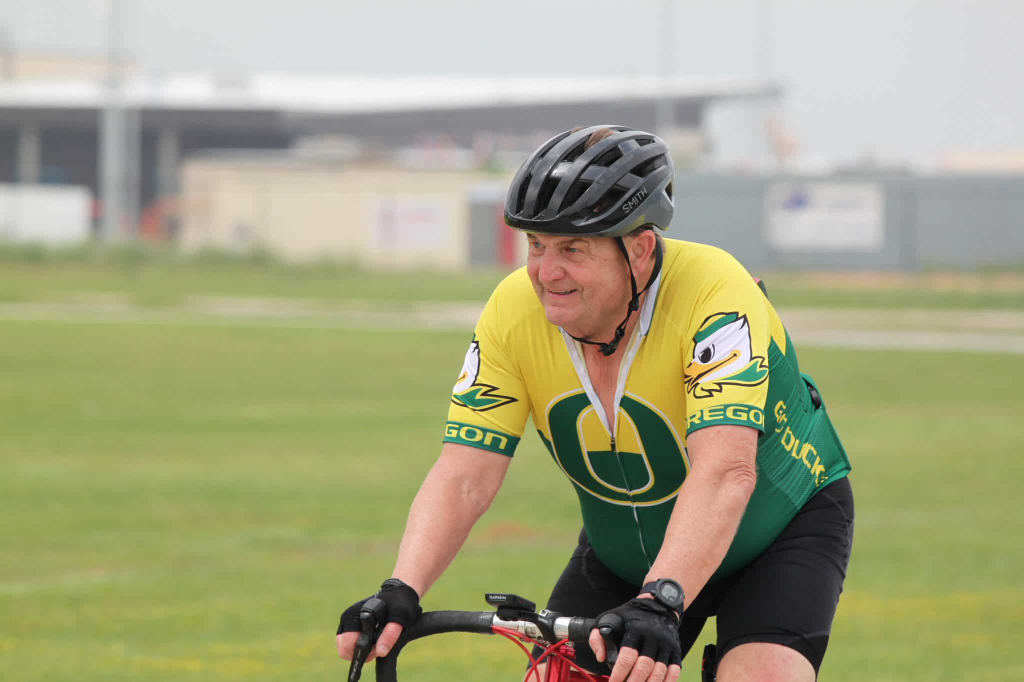 High-angle shot of a smiling cyclist wearing a Green Oregon Ducks jersey and helmet, riding outdoors on a cloudy day.