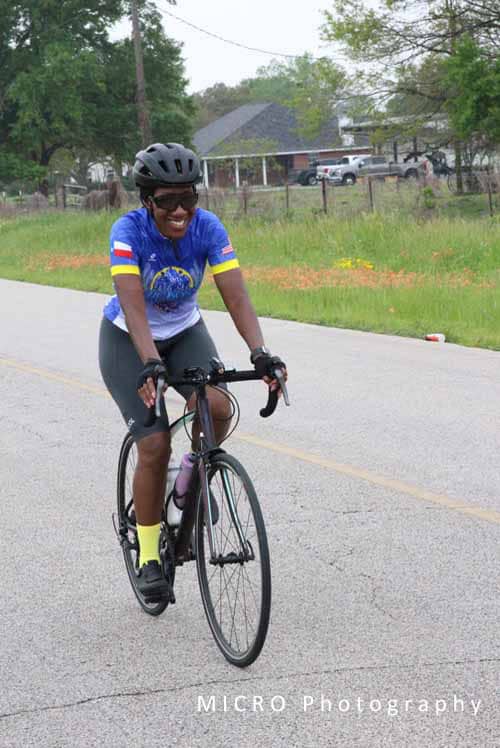 Smiling woman cycling on road with green trees and houses in background.