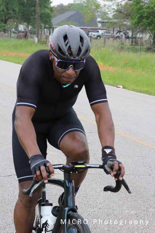 1. Male cyclist riding a bike on a road with greenery in the background, wearing a helmet and sunglasses.