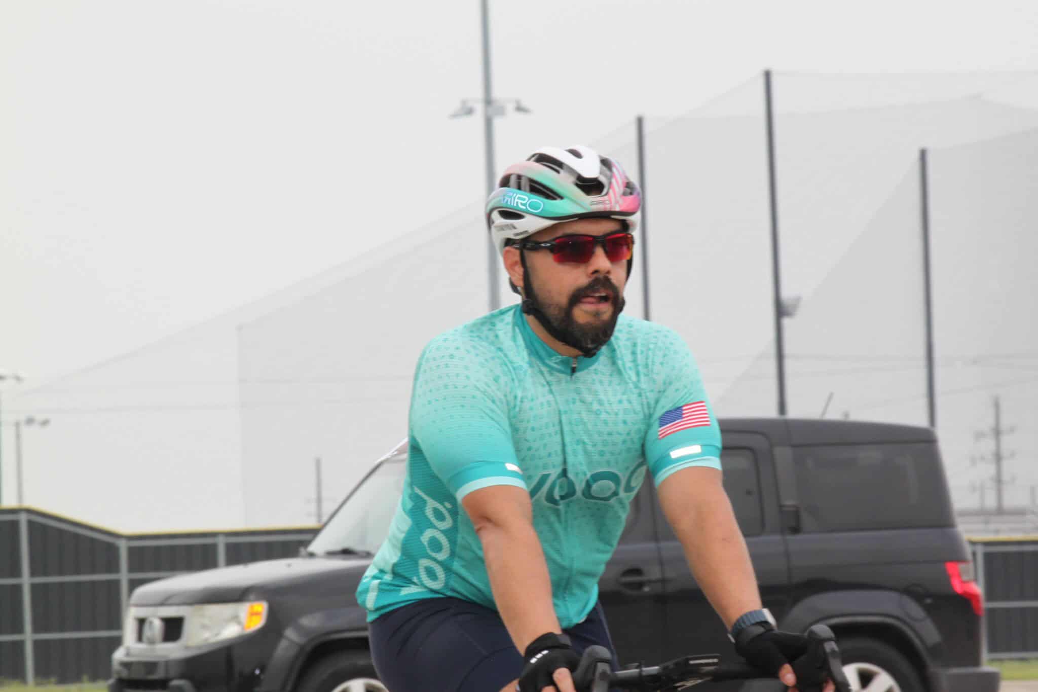 Vintage-style race cyclist on a blue road bike, wearing a helmet and sunglasses, at Bluebonnet Express event.