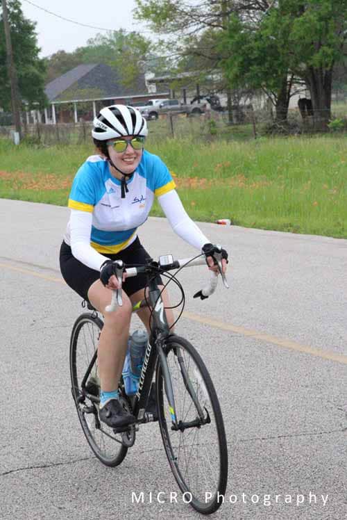 Smiling woman riding a bike on a paved road during daytime, wearing a helmet and sunglasses, promoting outdoor cycling.