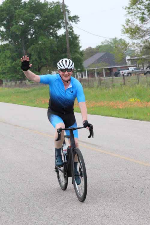 Smiling cyclist riding a blue bike on the road, wearing a helmet and cycling gear.