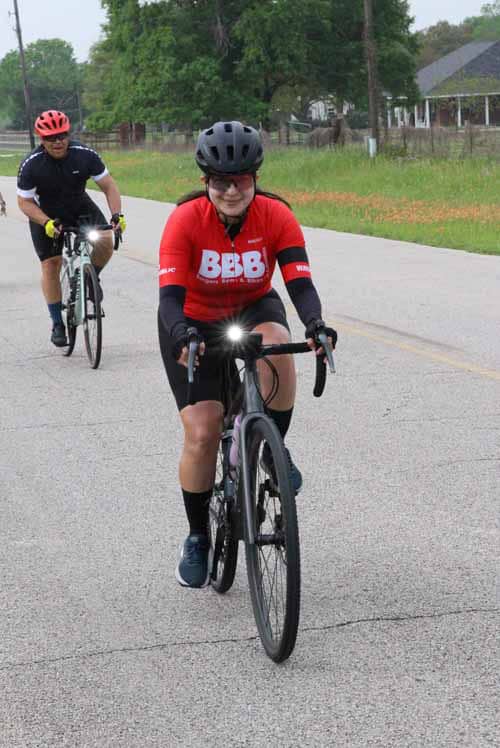 - Female cyclist riding on a rural road during daytime, wearing a red BBB jersey and helmet for Bluebonnet Express.