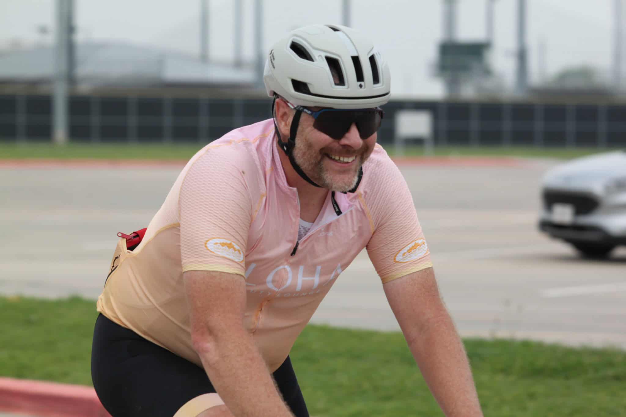 Cyclist riding on a bike, wearing helmet and sunglasses, smiling outdoors at Bluebonnet Express.