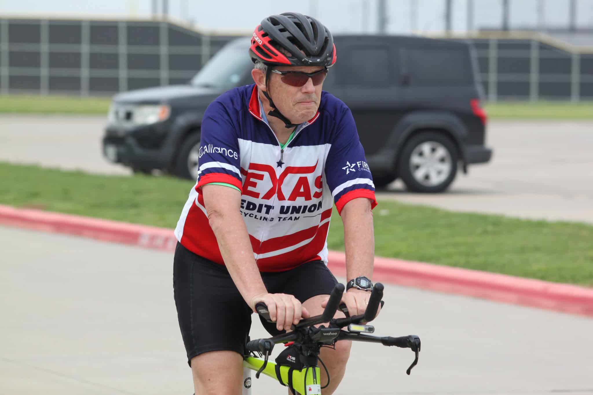 Active cyclist wearing Texas Credit Union cycling team jersey, helmet, and sunglasses riding a bike outdoors.