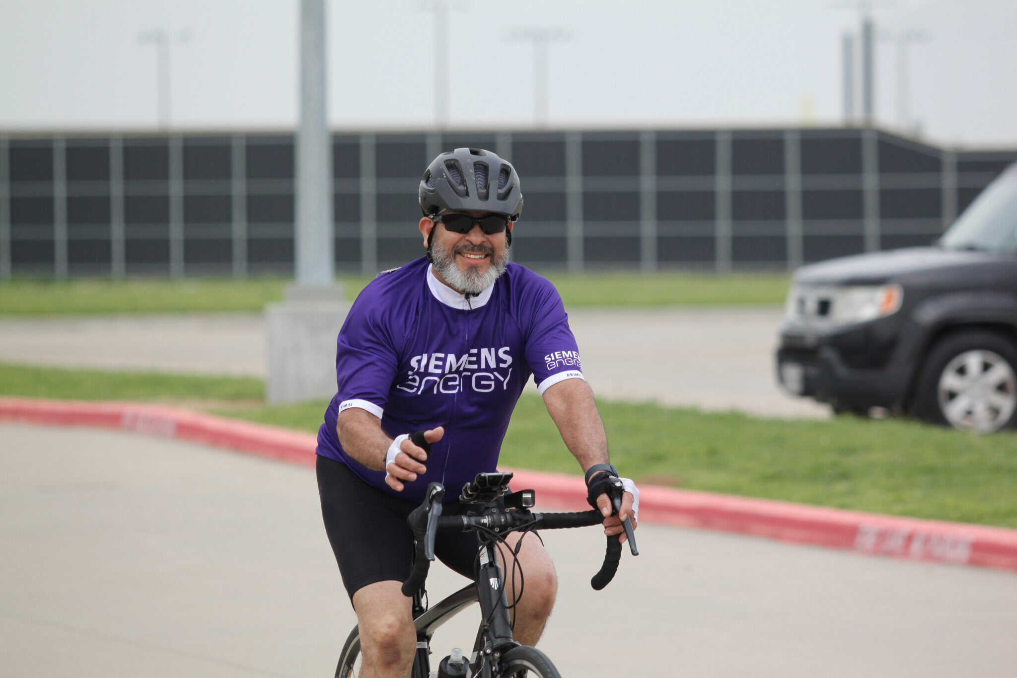 Smiling man cycling outdoors in purple Siemens Energy jersey, wearing helmet and sunglasses.