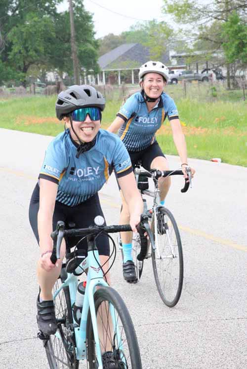 Smiling women cycling on a scenic road, promoting Bluebonnet Express bike rides and outdoor fun.