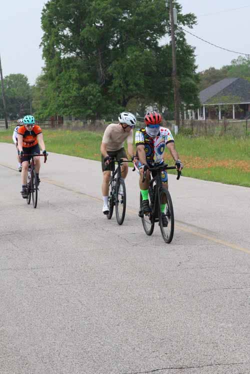 Two cyclists riding on a rural road during daytime with greenery and houses in the background.