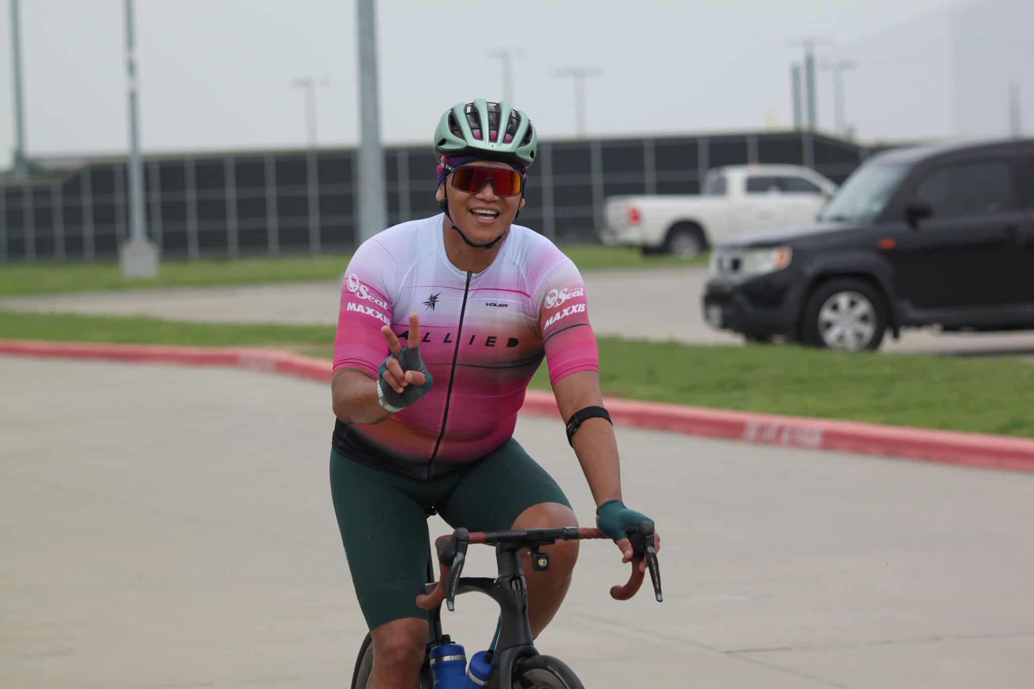 Bright female cyclist wearing a pink and white jersey, helmet, and sunglasses, riding a road bike outdoors.