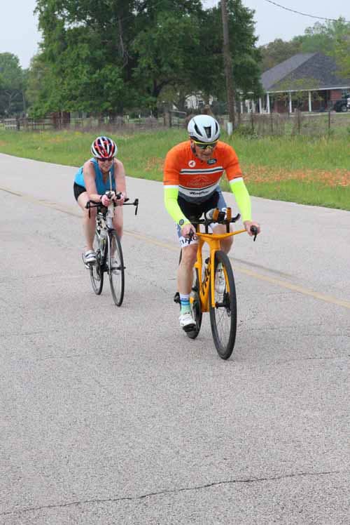 Bright orange and blue cycling jerseys, two cyclists riding on a rural road during daytime.
