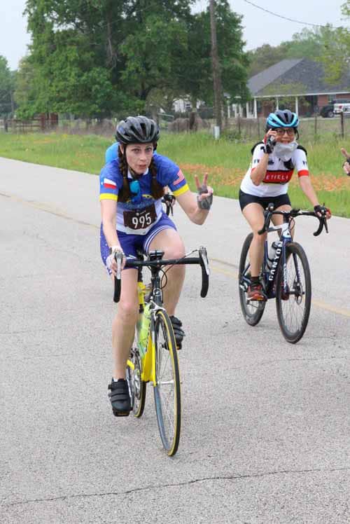 Bright female cyclist in colorful jersey during Bluebonnet Express race, riding on rural road with supportive friends.
