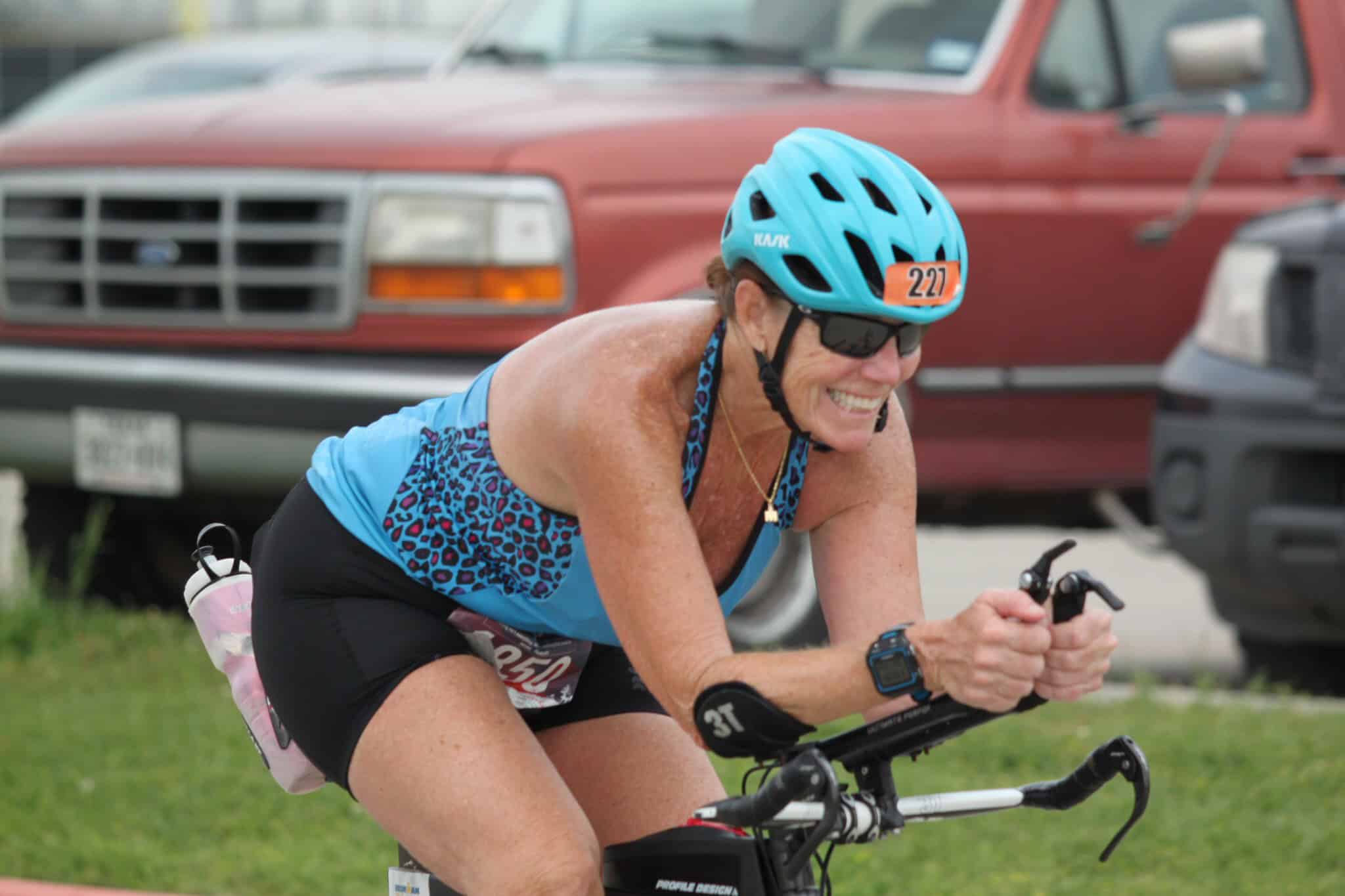 Bright blue cycling helmet and race bib, smiling female athlete riding a bike outdoors for Bluebonnet Express.