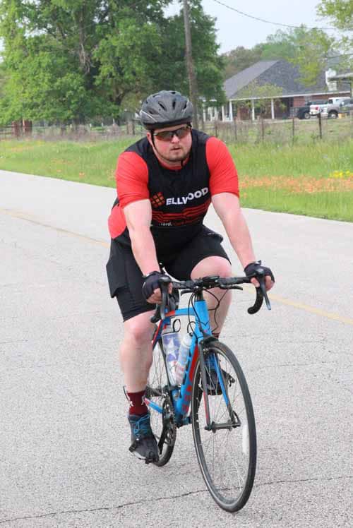 Bike rider wearing a red and black shirt, helmet, cycling on a rural road.