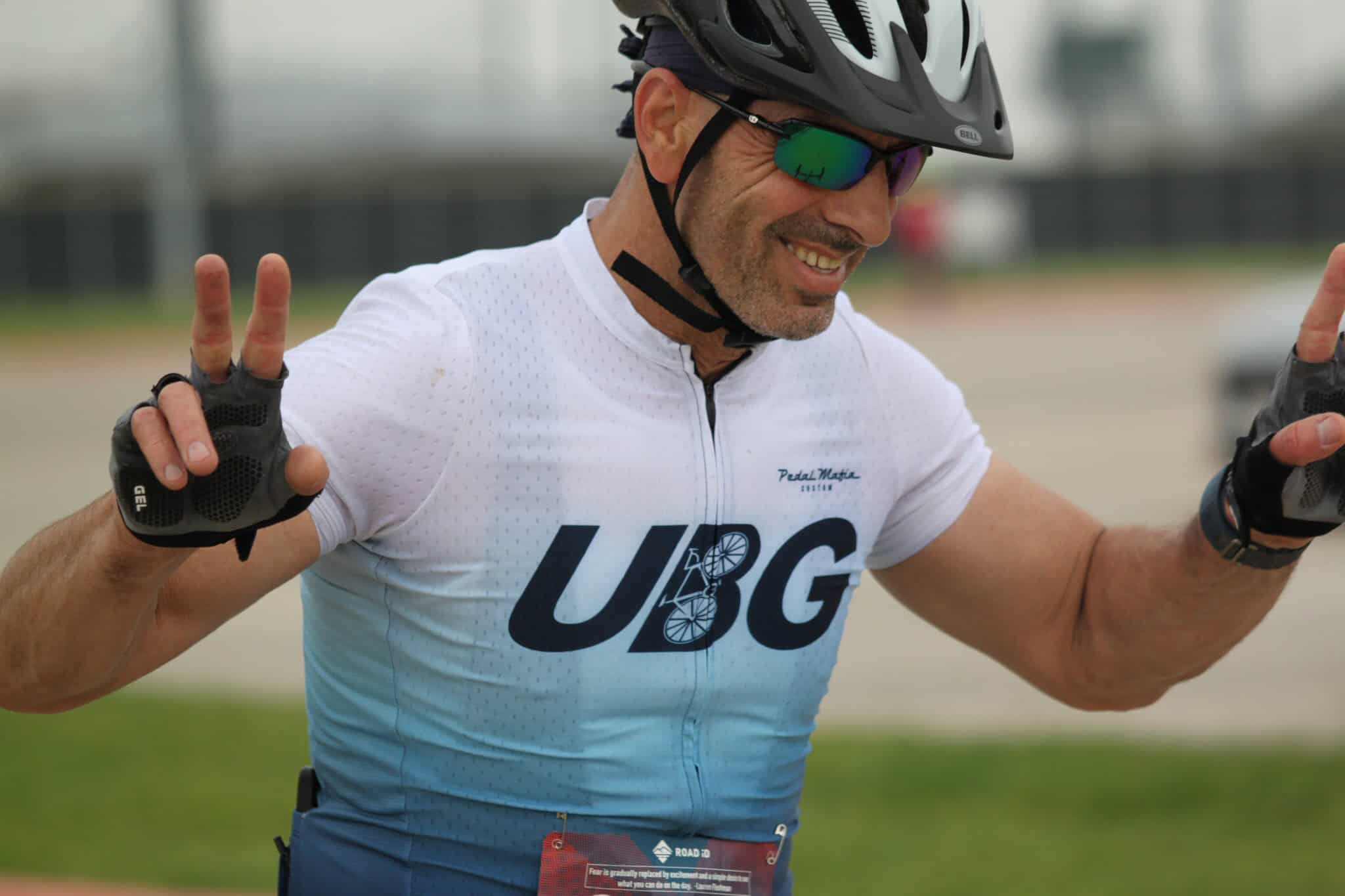 Vibrant cyclist wearing UBG gear, smiling, making peace signs after ride, outdoor biking event, road cycling, bike enthusiast, energetic sport.
