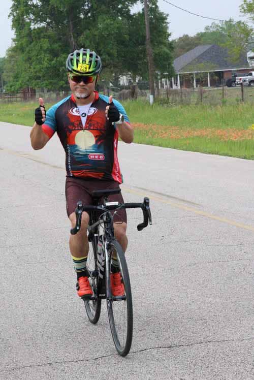 Brightly dressed cyclist riding on a suburban road with thumbs up gesture, promoting Bluebonnet Express bike rides.