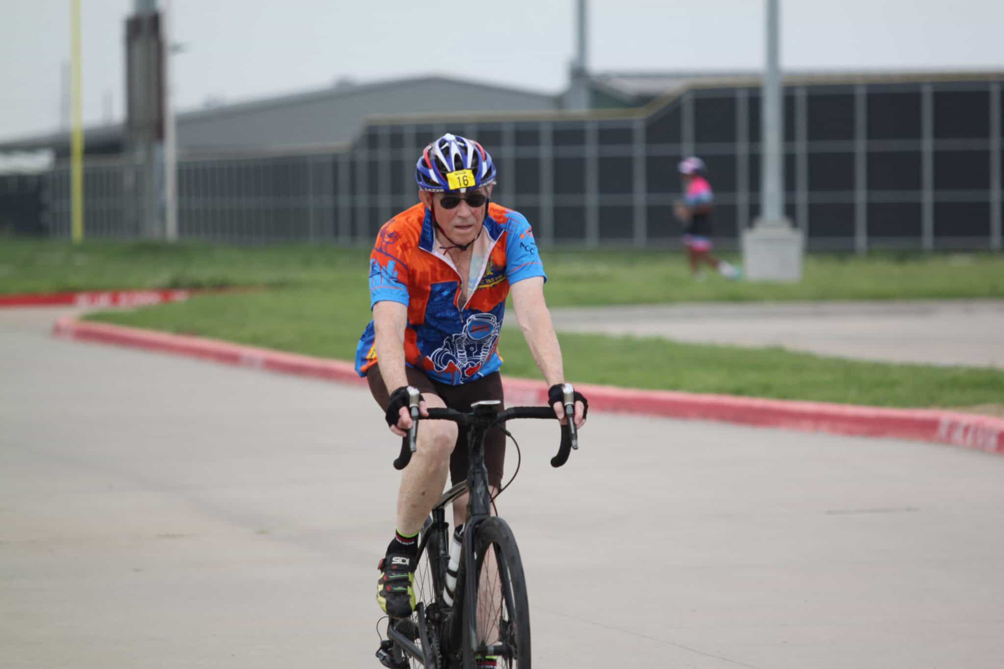 Older male cyclist racing on a bike during an event, wearing a colorful jersey and helmet.