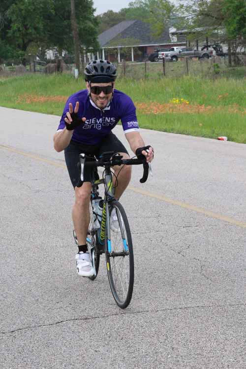 Bright cyclist wearing a blue jersey, riding a road bike on a rural route, smiling and making a peace sign.