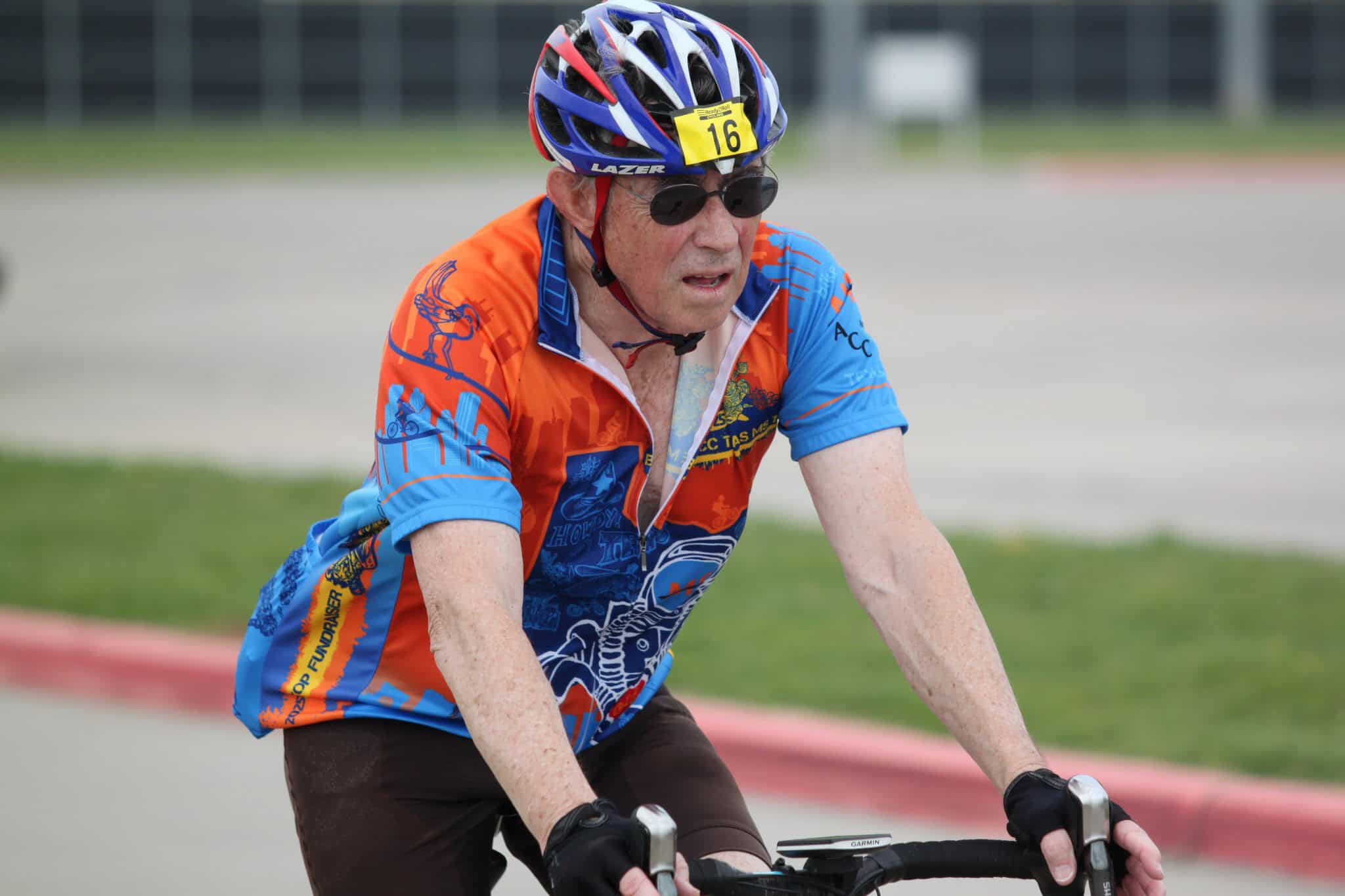 Elderly man cycling in a race, wearing a blue and orange jersey and helmet.