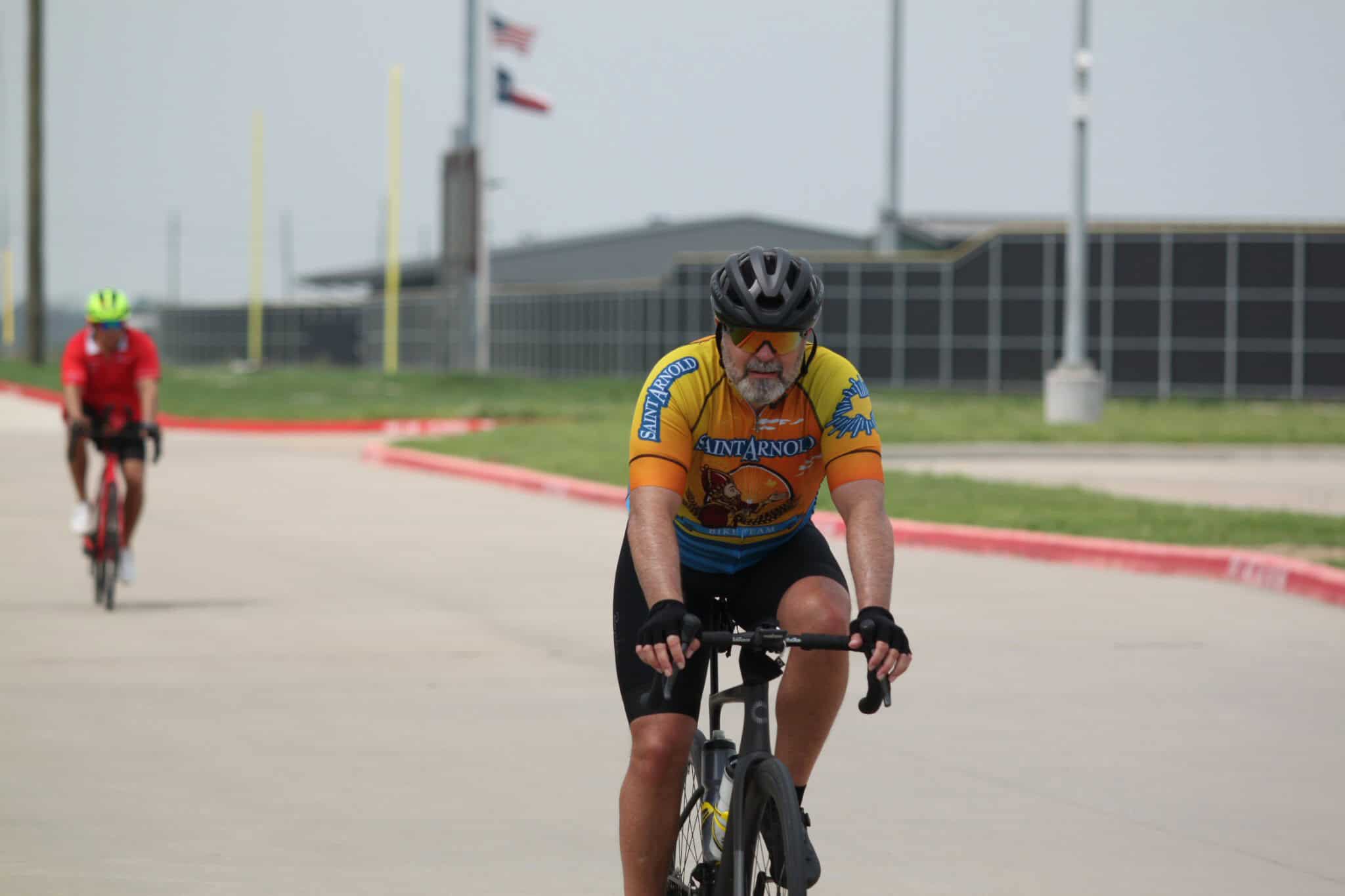 Bright man cycling outdoors wearing Saint Arnold race jersey and helmet, participating in a bike event.
