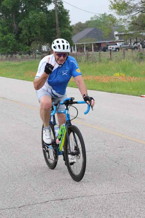 Happy cyclist riding a blue bike on the road, smiling and waving at the camera.