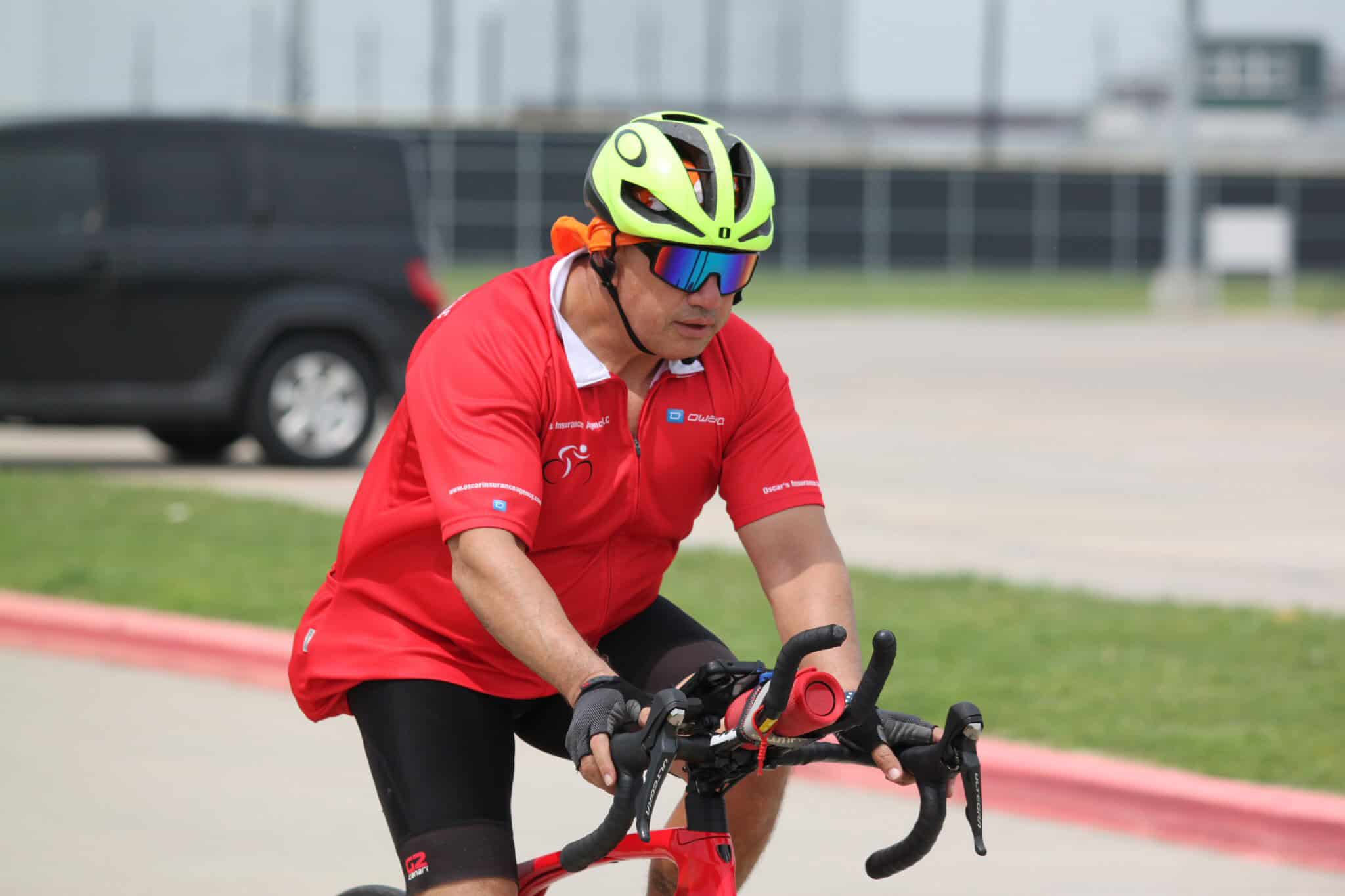 Helmeted man riding a bicycle with safety gear, outdoors, near roadside.