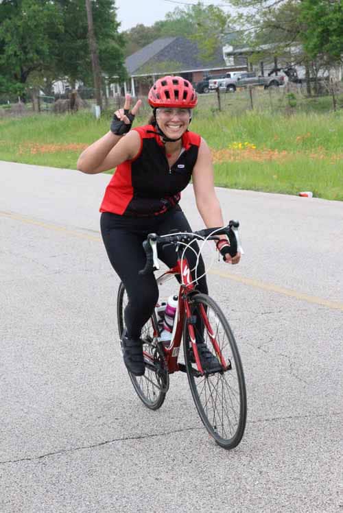 Cheerful cyclist wearing a red helmet and sports attire riding on rural road, smiling and making victory sign.