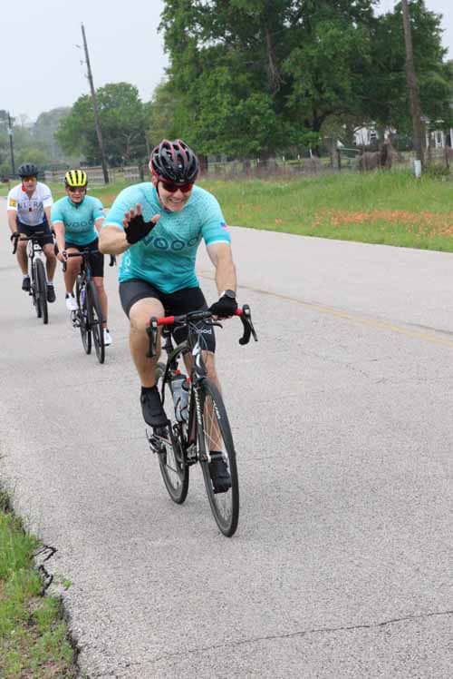 Smiling cyclists riding together on a scenic rural road during daytime, promoting outdoor activity and community spirit.
