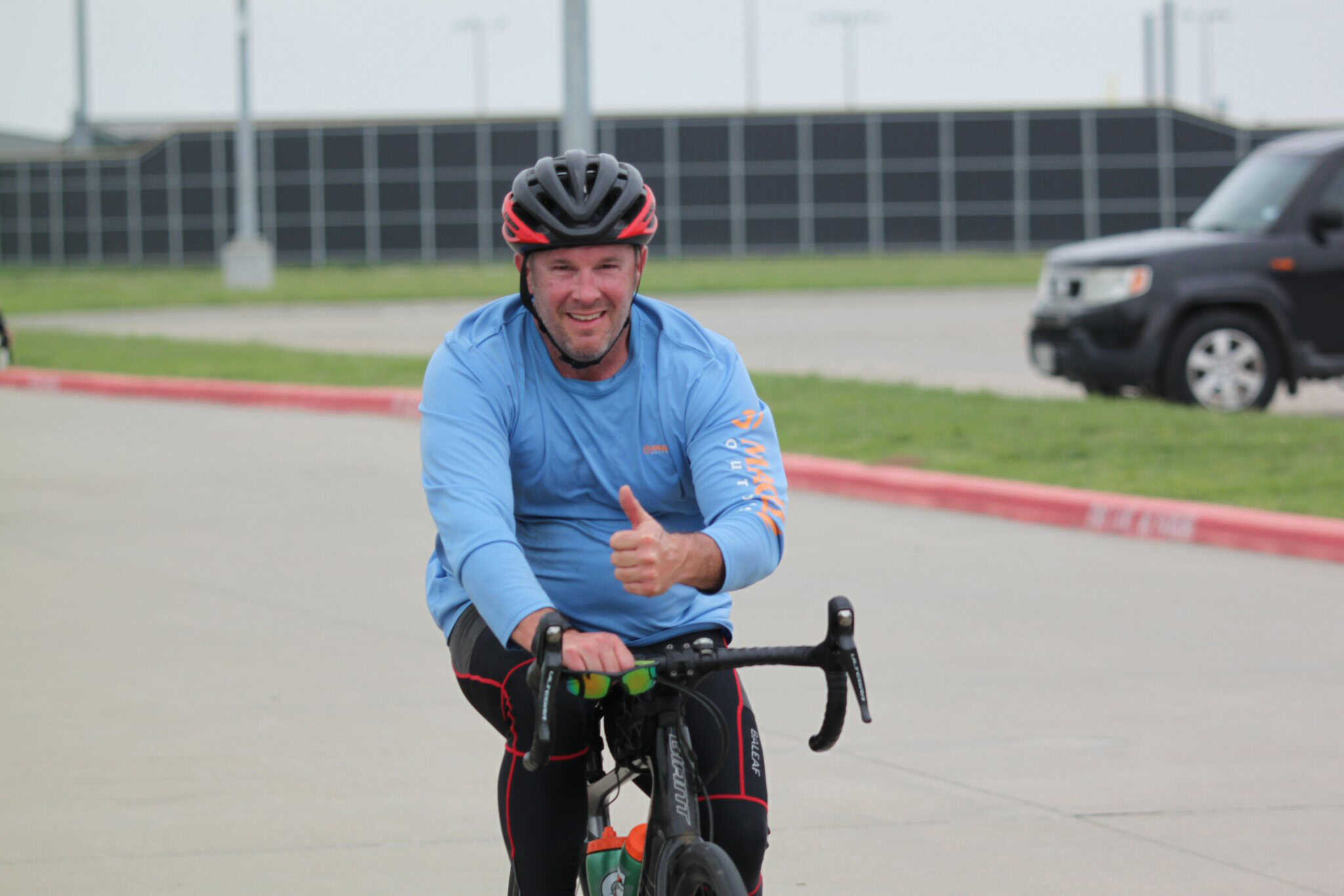 Happy male cyclist with thumbs up riding on a road, wearing helmet and sportswear, outdoors.