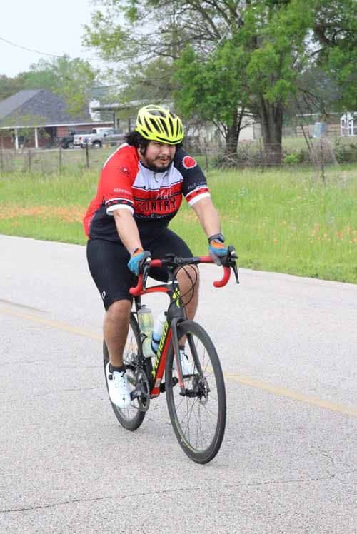 Bicycle rider wearing a yellow helmet, red and black cycling jersey on a suburban road.