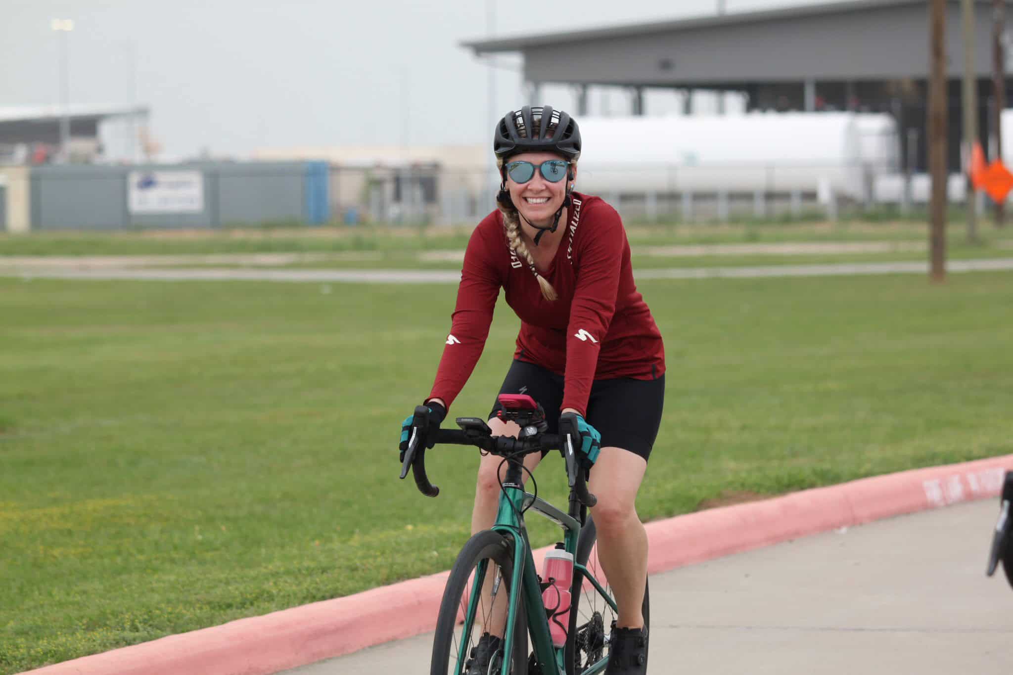 Bright woman cyclist smiles on green bike, wearing helmet, sunglasses; enjoying outdoor Bluebonnet Express ride.