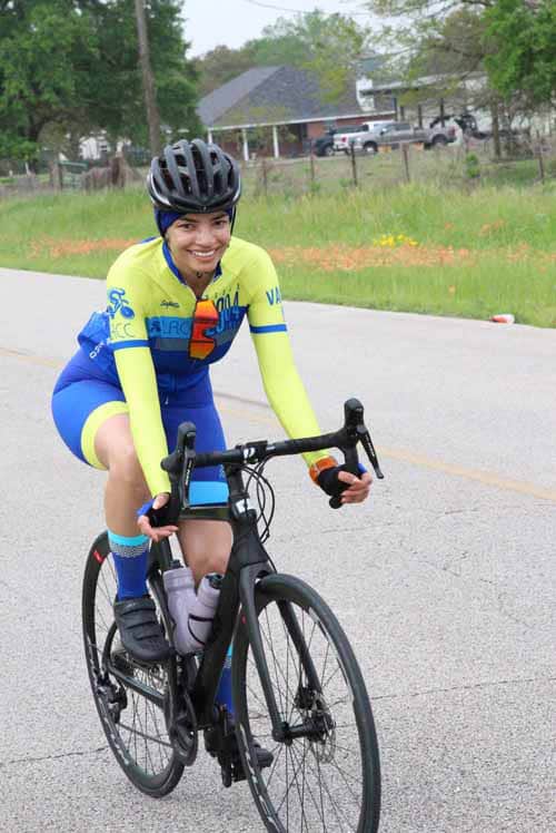 Bright smiling cyclist riding a black road bike in vibrant blue and yellow jersey outdoors.