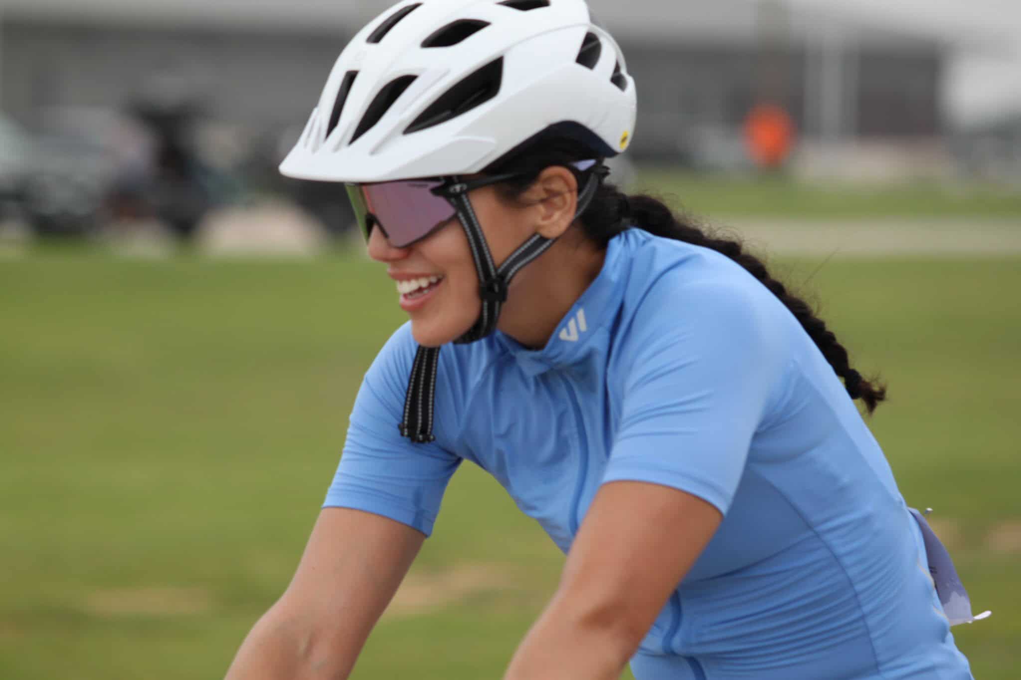 Bright female cyclist smiling, wearing a helmet and sunglasses, riding a blue bike on green outdoor trail.
