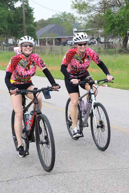 Two women riding bikes in colorful jerseys during a sunny outdoor event.