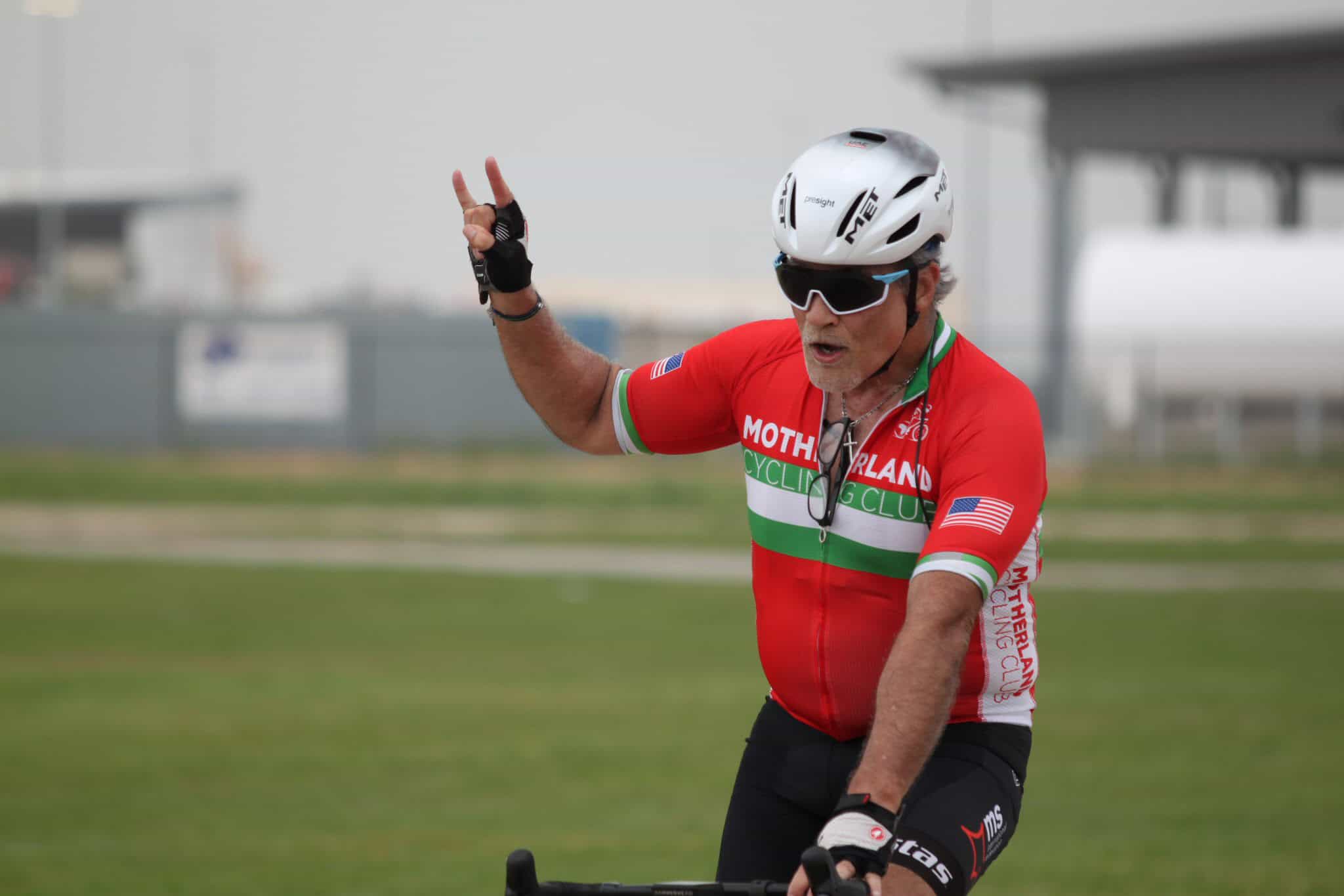 Older male cyclist with helmet and sunglasses, making peace sign during a ride, showcasing active biking lifestyle.