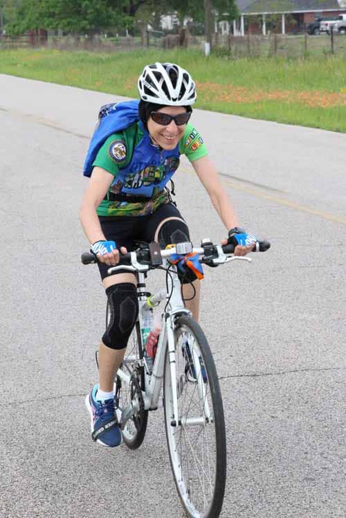 Bright woman cycling on road, wearing safety gear, smiling, in an outdoor park setting, promoting active lifestyle and biking safety.