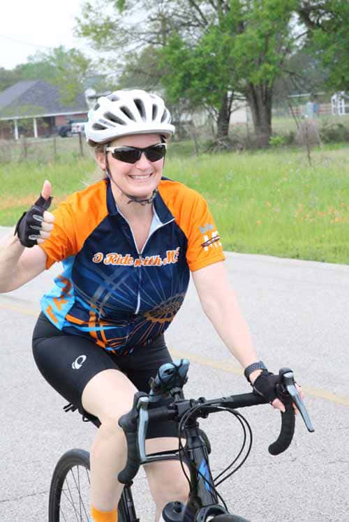 Happy female cyclist wearing a Bluebonnet Express jersey, riding a bike outdoors on a scenic route.