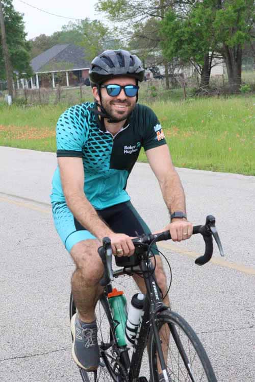 Bright smiling cyclist riding road bike wearing blue jersey and helmet, outdoor scenic route, promoting Bluebonnet Express.