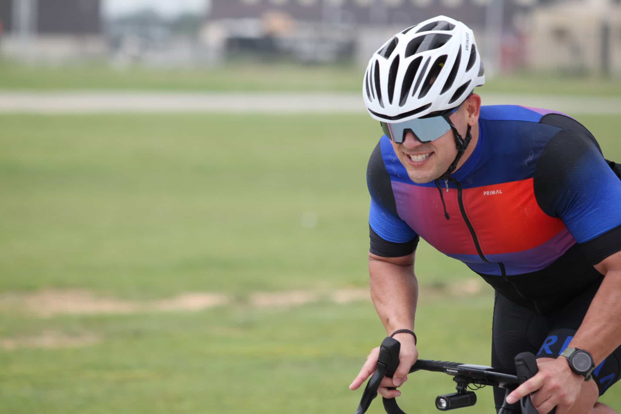 Bright male cyclist in blue and red cycling jersey, wearing a helmet and sunglasses, riding on a grassy trail.